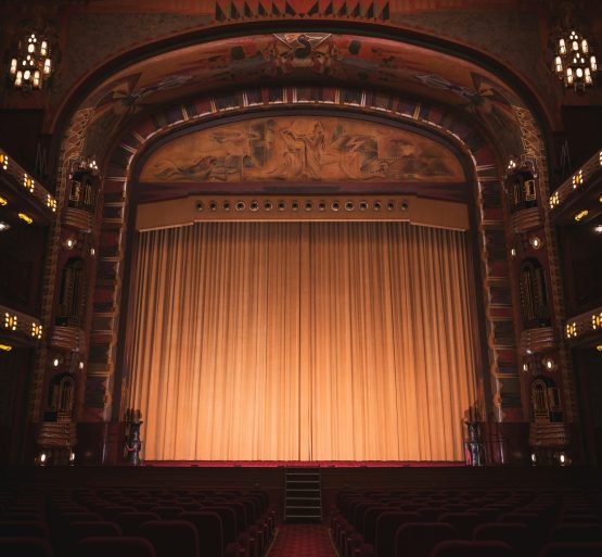 Image shows an old style theatre stage, with a closed curtain lit from below.