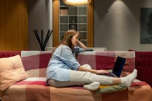 An Asian woman is sitting comfortably on a sofa covered in a tartan blanket. The woman is smiling softly with one hand on her laptop.