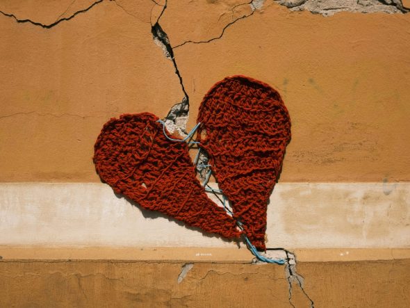 The image shows a cracked orange wall behind a broken red heart. The two pieces of the heart have been repaired with wire.