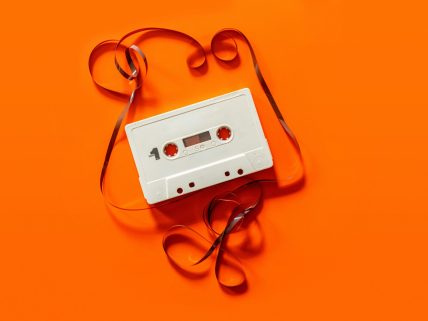 A white cassette tape with its side facing up, labelled 'Side 1', unspooled tape looping and curling decoratively around it, set against a bright orange background.